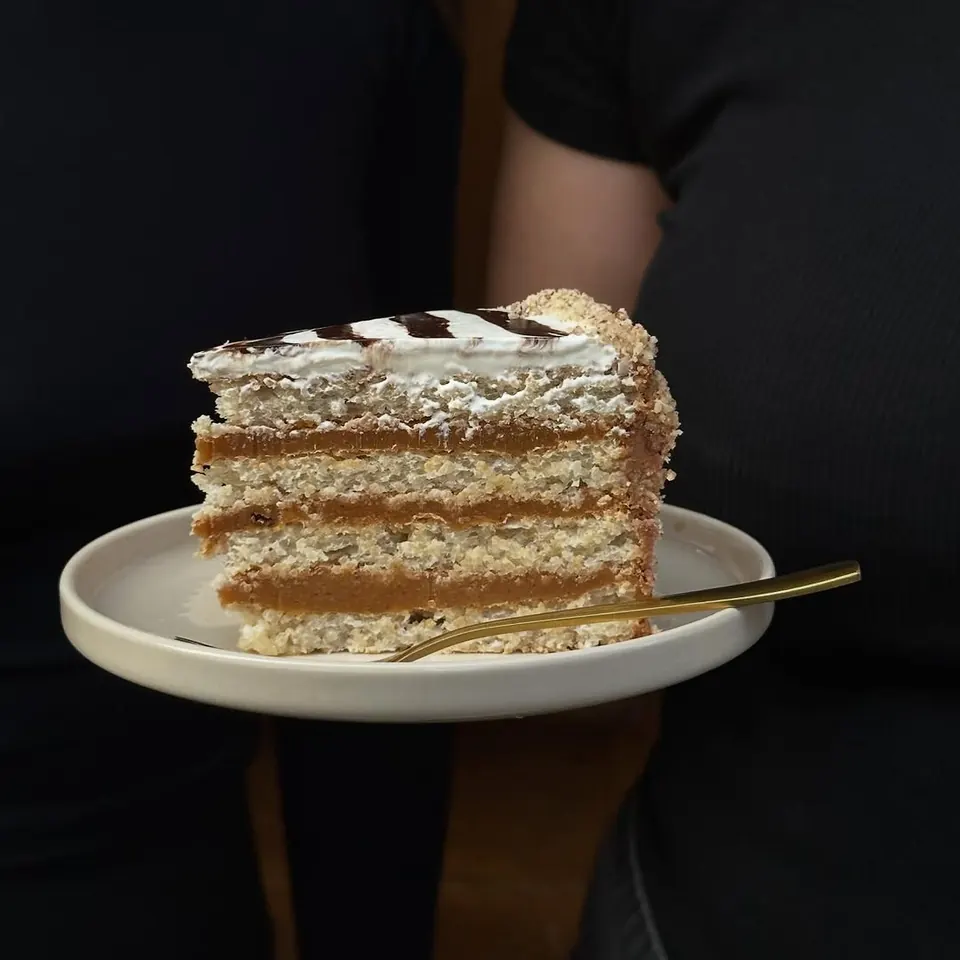 A tall slice of Esterházy cake with caramel layers on a cream plate with a gold fork